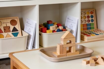 Educational toy sets stored separately in a cupboard beside an unfinished block structure on a dining-table tray.