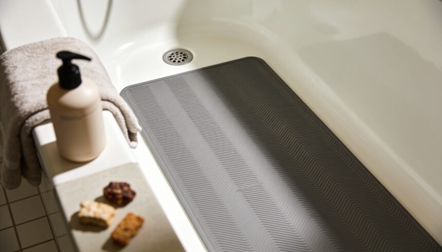 An empty bath with a rubber non-slip mat in the bottom, a folded towel and an unlabelled bottle on the edge.