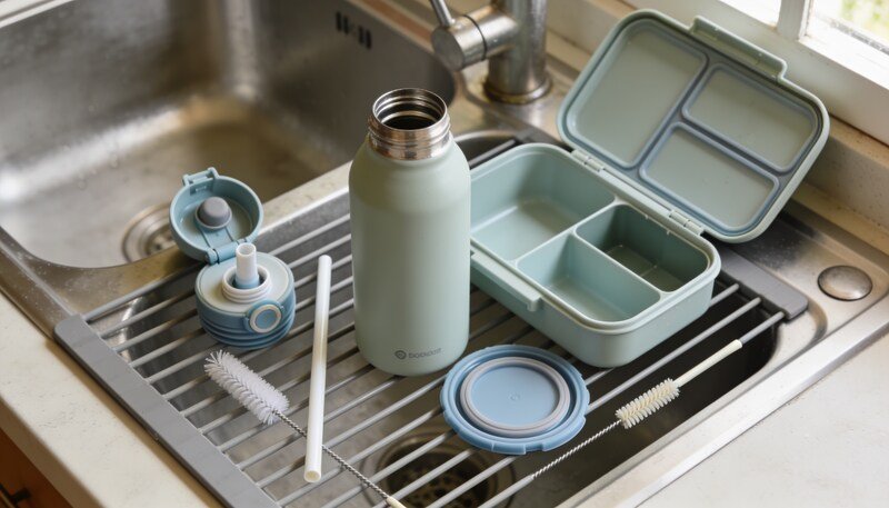 Dismantled drink bottle and lunchbox with separate straws, lids and silicone seals drying beside cleaning brushes.