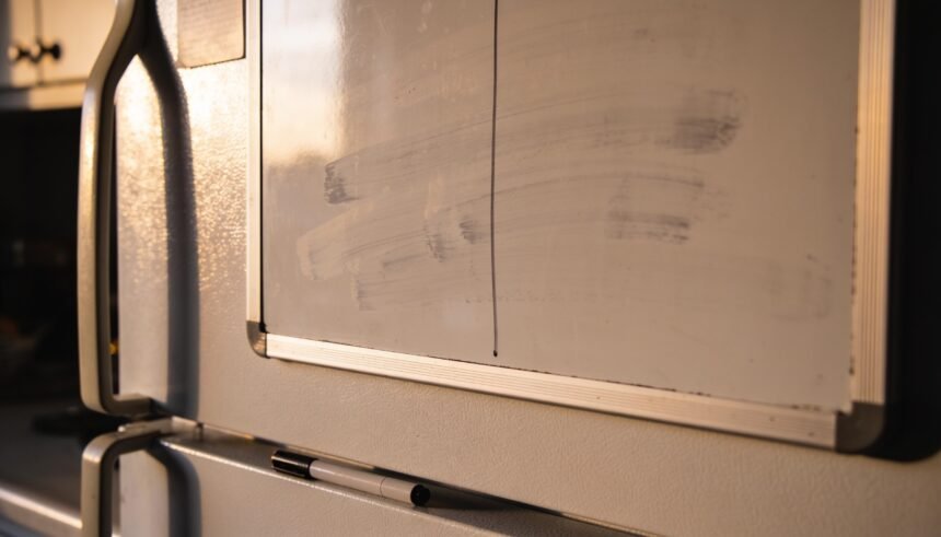 A fridge whiteboard wiped almost clean with a marker lying on the bench below, lid off.