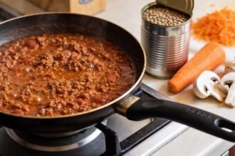 A pan of bolognese on a stovetop beside a tin of lentils, grated carrot and mushrooms on the bench.