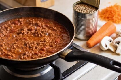 A pan of bolognese on a stovetop beside a tin of lentils, grated carrot and mushrooms on the bench.