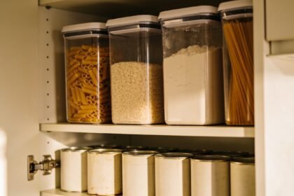 An open pantry with sealed clear containers of pasta, rice and flour above a row of plain tins.