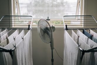 Two clothes airers with washing hung spaced apart, a pedestal fan pointed at them and an open window behind.