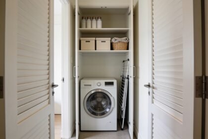 Open European laundry cupboard with a front loader, a shelf of plain boxes above it, unmarked bottles on a high shelf and a folded airer beside the machine.