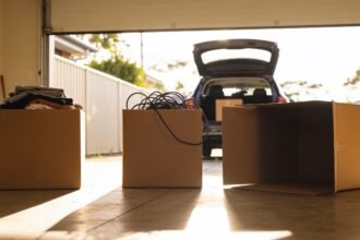 Three cardboard boxes on a garage floor holding folded clothes and tangled cables, with an open car boot behind.
