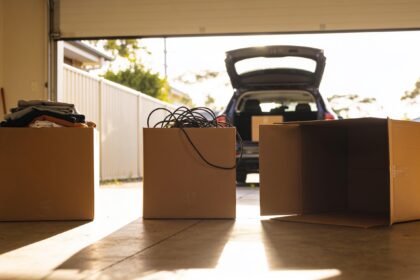 Three cardboard boxes on a garage floor holding folded clothes and tangled cables, with an open car boot behind.