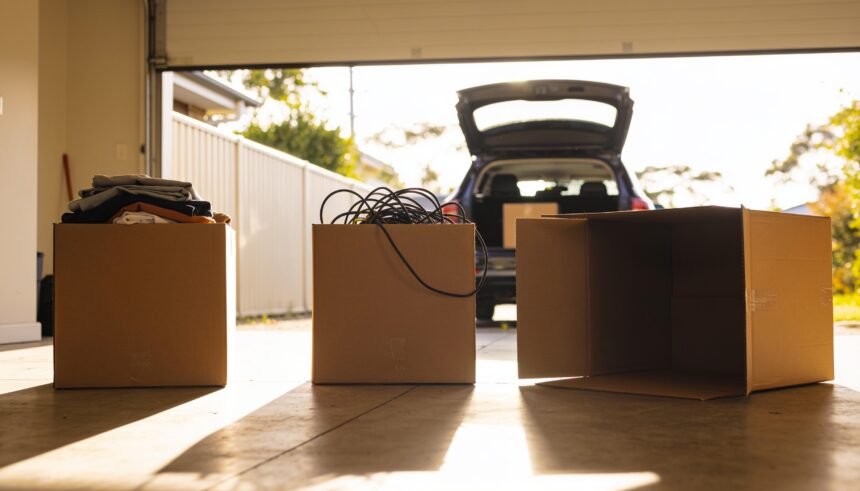 Three cardboard boxes on a garage floor holding folded clothes and tangled cables, with an open car boot behind.
