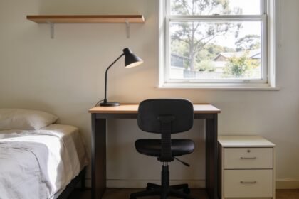 A narrow study desk under a bedroom window with an adjustable chair, desk lamp and a shelf above.