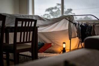 A sheet fort built over dining chairs and a clothes airer in a lounge room with rain on the window behind.