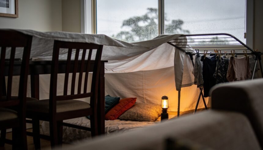A sheet fort built over dining chairs and a clothes airer in a lounge room with rain on the window behind.