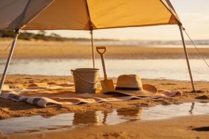 A beach shelter and towel set up on sand with a bucket, spade and wide-brim hat beside them at low tide.