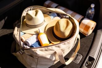 An open bag in a car boot packed with towels, two broad-brim hats, sunscreen, a picnic rug and a water bottle.