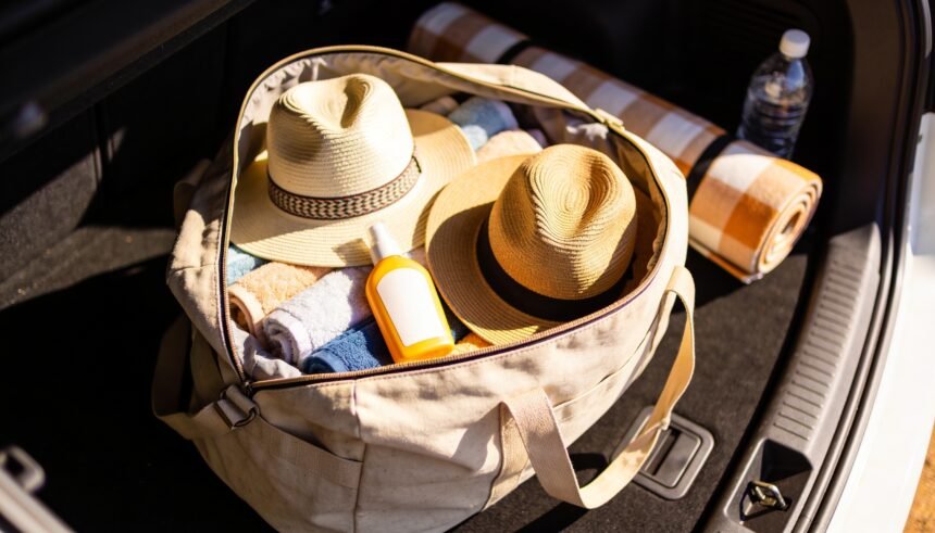 An open bag in a car boot packed with towels, two broad-brim hats, sunscreen, a picnic rug and a water bottle.