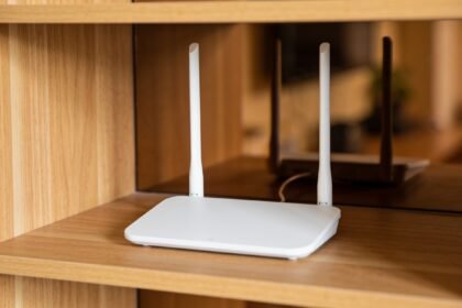 A plain white wireless router standing in the open on a timber shelf, clear of surrounding objects.