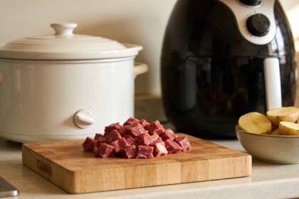 A slow cooker and an air fryer side by side on a bench with diced beef and halved potatoes between them.