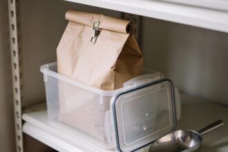A rolled and clipped pet food bag standing inside an open airtight container on a pantry shelf, with a scoop beside it.