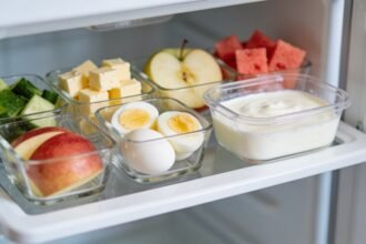 A fridge shelf at child height stocked with clear glass containers of cut vegetables, cheese, boiled eggs, fruit and yoghurt.