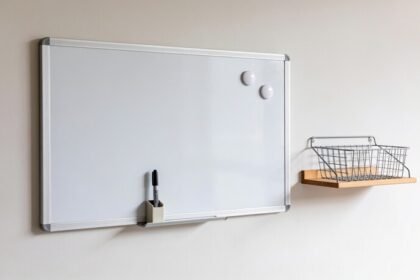 A blank white magnetic whiteboard on a pale wall beside a small shelf and wire basket, with a marker in a holder on its lower edge.