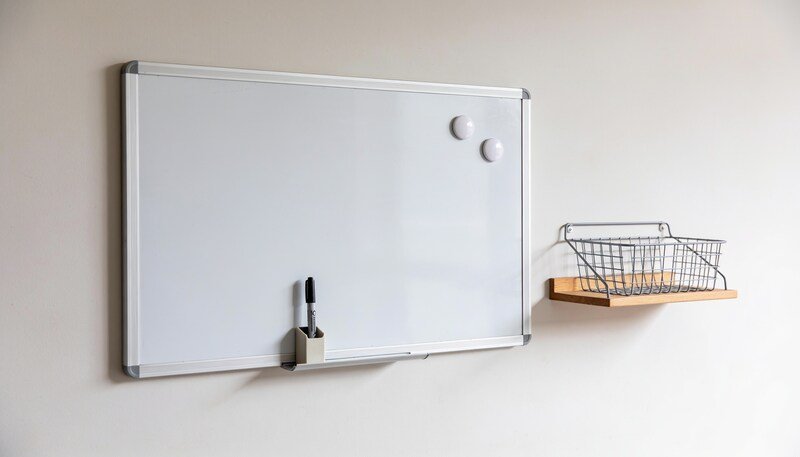 A blank white magnetic whiteboard on a pale wall beside a small shelf and wire basket, with a marker in a holder on its lower edge.