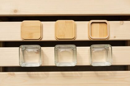 Three plain bamboo container lids laid flat in a row above three empty square glass container bases on a timber bench, seen from directly above.
