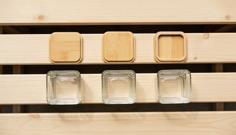 Three plain bamboo container lids laid flat in a row above three empty square glass container bases on a timber bench, seen from directly above.