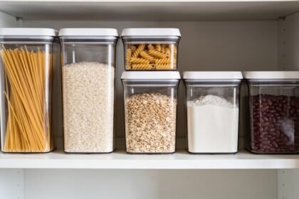Six clear square pantry containers of different heights on a white shelf, filled with pasta, rice, oats, flour and dried beans.