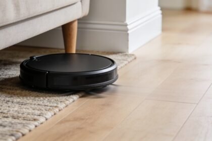 A round black robot vacuum on timber floorboards passing the edge of a flat-weave rug beside a sofa leg.