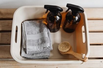 Looking down into a plain cleaning caddy on a timber bench holding two amber glass spray bottles, folded grey microfibre cloths and a scrubbing brush.