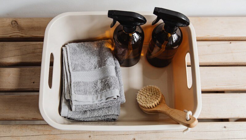 Looking down into a plain cleaning caddy on a timber bench holding two amber glass spray bottles, folded grey microfibre cloths and a scrubbing brush.