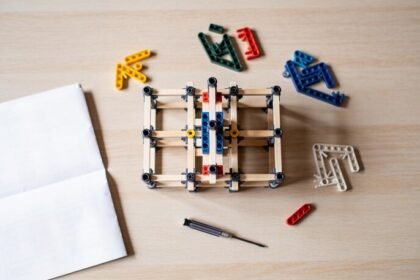 A partly assembled construction toy on a timber table surrounded by loose coloured building pieces, a small screwdriver and a folded paper sheet.