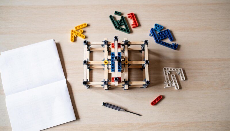 A partly assembled construction toy on a timber table surrounded by loose coloured building pieces, a small screwdriver and a folded paper sheet.