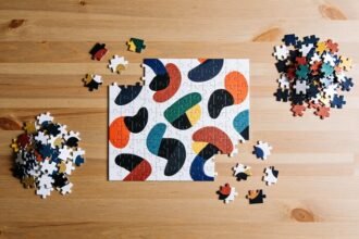 A part-completed jigsaw puzzle of coloured abstract shapes on a timber table, with loose pieces sorted into two small piles beside it.