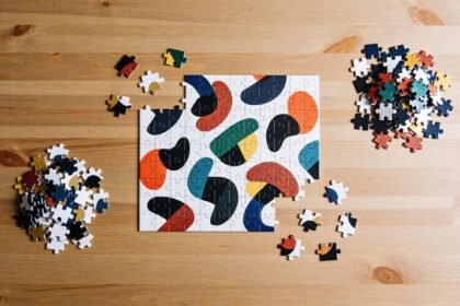 A part-completed jigsaw puzzle of coloured abstract shapes on a timber table, with loose pieces sorted into two small piles beside it.