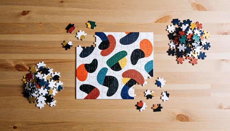 A part-completed jigsaw puzzle of coloured abstract shapes on a timber table, with loose pieces sorted into two small piles beside it.