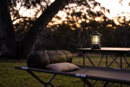 A camp stretcher on grass beside a folding table at dusk, with a rolled sleeping bag and pillow on the stretcher and a lit lantern on the table.