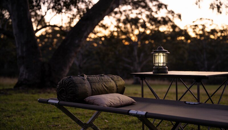 A camp stretcher on grass beside a folding table at dusk, with a rolled sleeping bag and pillow on the stretcher and a lit lantern on the table.