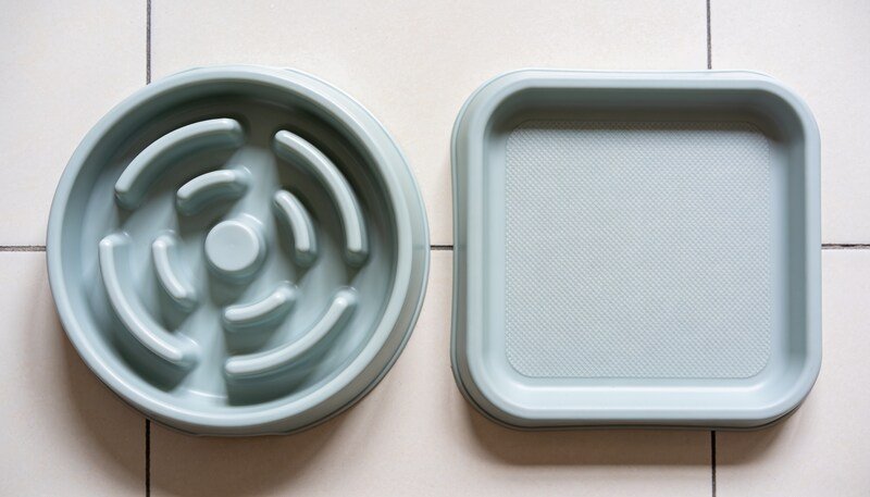 An empty round maze feeding bowl with raised ridges beside a flat square textured silicone lick mat on pale tiles.