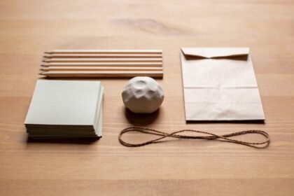 A stack of plain paper squares, a row of wooden pencils, a ball of pale clay, a paper bag and brown string arranged on a timber table.