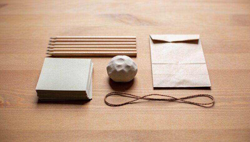 A stack of plain paper squares, a row of wooden pencils, a ball of pale clay, a paper bag and brown string arranged on a timber table.