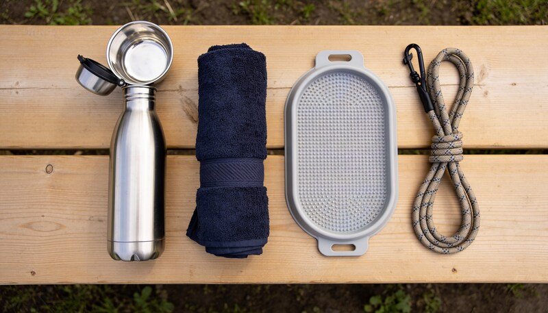 A stainless steel dog drink bottle with a fold-out bowl, a rolled navy towel, a silicone lick mat and a coiled rope lead on a timber bench.