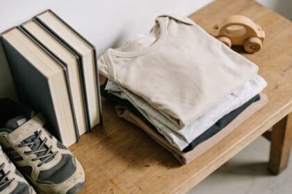 A folded pile of children's clothes on a timber table beside worn sports shoes, three books standing with their page edges to the camera, and a small wooden toy car.