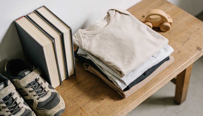 A folded pile of children's clothes on a timber table beside worn sports shoes, three books standing with their page edges to the camera, and a small wooden toy car.