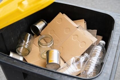 Looking straight down into an open recycling bin under its raised yellow lid, holding loose flattened cardboard, plain glass jars, aluminium cans and plastic bottles, none of it bagged.