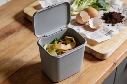 An open grey kitchen caddy on a timber bench holding vegetable peelings, with egg shells, coffee grounds and sheets of newspaper on a chopping board beside it.