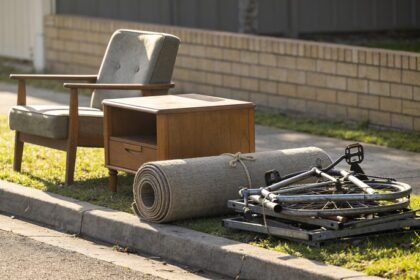 A hard rubbish pile sorted into three groups on a nature strip: an armchair and side table, a rolled tied carpet, and stacked scrap metal with a bike frame.
