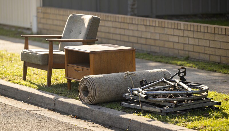 A hard rubbish pile sorted into three groups on a nature strip: an armchair and side table, a rolled tied carpet, and stacked scrap metal with a bike frame.