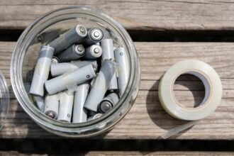 A clear lidded jar on a timber bench half full of used batteries with clear tape across their ends, and a roll of tape beside it.