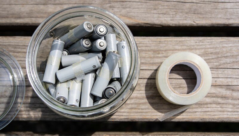 A clear lidded jar on a timber bench half full of used batteries with clear tape across their ends, and a roll of tape beside it.