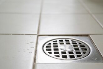 A stainless steel floor waste drain set into pale tiles, photographed close to floor level with water beading on the tiles beside it.